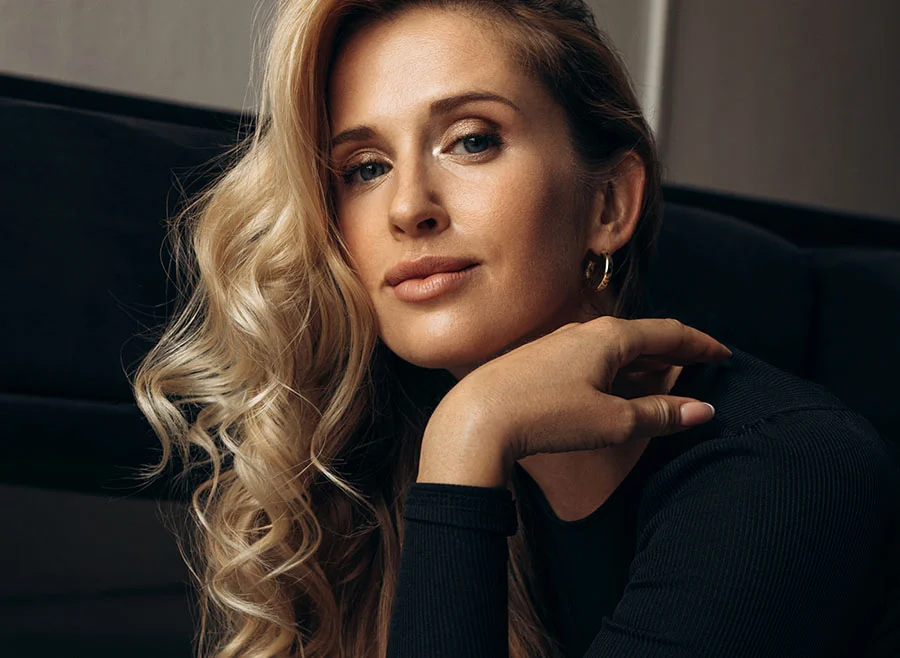 A blonde woman with soft curls wears a black top and rests her chin on her hand - Dermal Fillers in Frisco, TX