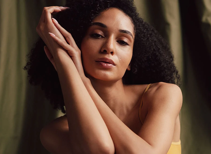 A woman with natural curls poses gracefully, wrapping her arms around her head - Forma in Frisco, TX