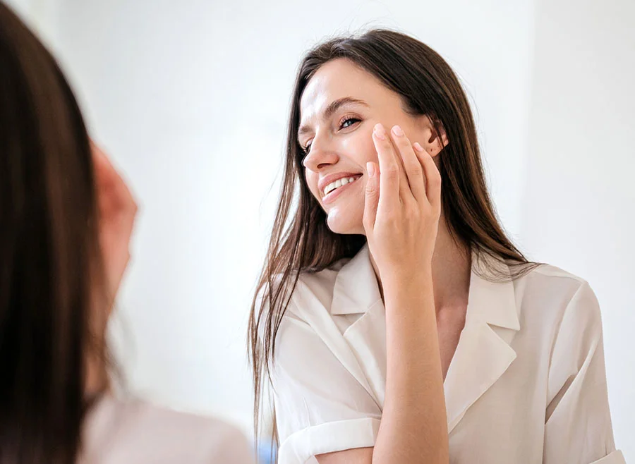 A smiling brunette woman with long straight hair applies skincare to her cheek while looking at herself in the mirror - Fraxel Laser in Frisco, TX