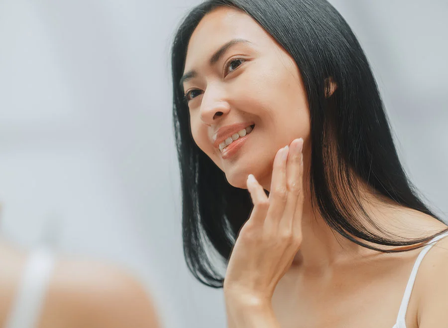 A woman with long, straight dark hair smiles as she gently touches her face while looking into the mirror - Skin Tightening in Frisco, TX