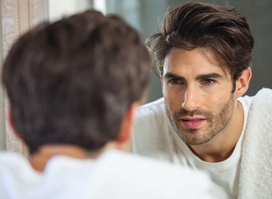 A man with styled brown hair looks seriously into the mirror, wearing a white t-shirt, with a focused and thoughtful expression - Tear Through Fillers in Frisco, TX